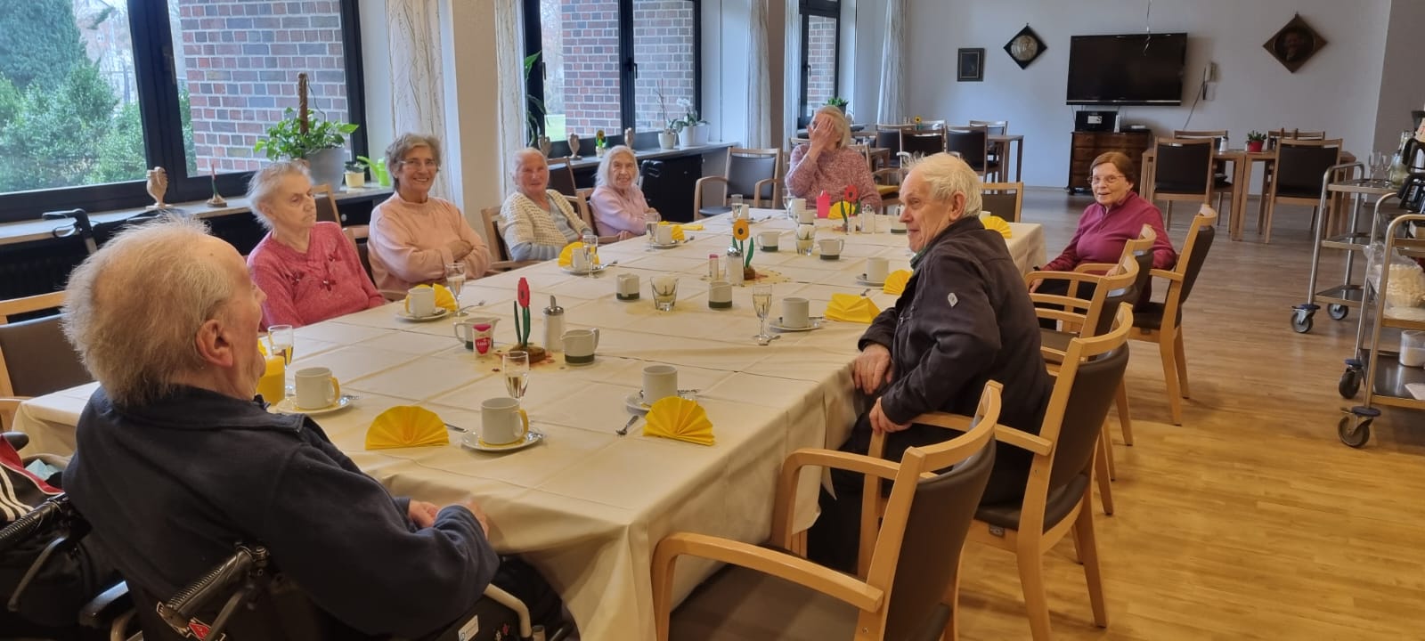Die Seniorinnen und Senioren gemeinsam am festlich gedeckten Kaffeetisch im Altenheim, während sie fröhlich auf die Geburtstagstorten warten.
