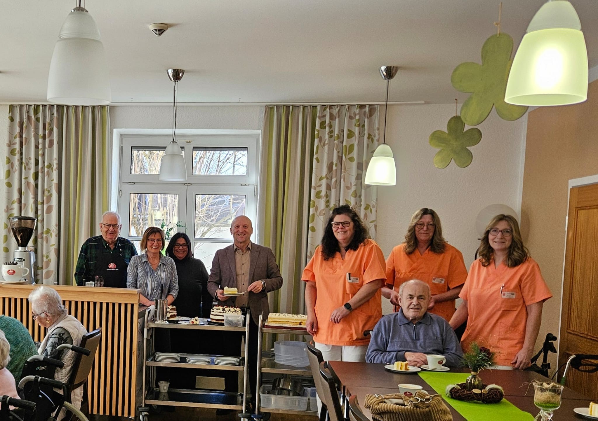 Pfleger und Bewohner an einem Tisch, dahinter 4 Personen, eine davon teilt eine Torte aus